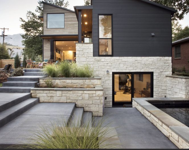 A modern multi-level house with dark siding and light-colored stone accents. Concrete stairs lead down to a lower patio area with a small pool or water feature and a door leading to a basement-level room. Lush green plants are integrated into the stone landscaping.